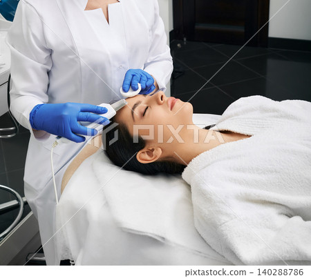 Ultrasound face peeling. Hispanic woman while ultrasonic peel forehead skin with skin scrubber at a beauty clinic Ultrasound face peeling. Hispanic woman while ultrasonic peel forehead skin with skin scrubber at a beauty clinic 140288786
