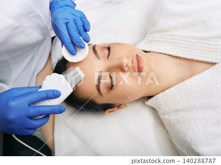 Ultrasound facial peeling. Brunette woman getting ultrasonic peel skin with skin scrubber at cosmetology clinic Ultrasound facial peeling. Brunette woman getting ultrasonic peel skin with skin scrubber at cosmetology clinic 140288787