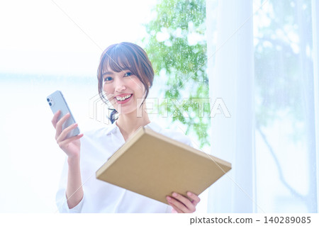 一位年輕女子微笑著站在窗邊,手裡拿著行李和智慧型手機。 一位年輕女子微笑著站在窗邊,手裡拿著行李和智慧型手機。 140289085