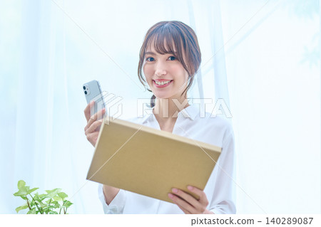 一位年輕女子站在明亮的窗邊微笑,手裡拿著她的隨身物品和智慧型手機。 一位年輕女子站在明亮的窗邊微笑,手裡拿著她的隨身物品和智慧型手機。 140289087