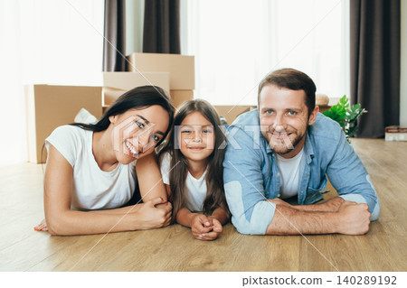 Happy family moving in new house. Mother, father and daughter together smiling lying down on floor with boxes on background Happy family moving in new house. Mother, father and daughter together smiling lying down on floor with boxes on background 140289192