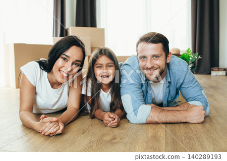Happy family moving in new house. Mother, father and daughter together smiling lying down on floor with boxes on background 140289193
