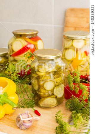 Jars filled with cucumbers, peppers, and zucchini sit on a wooden countertop. Fresh vegetables and herbs surround the jars as people prepare food for winter storage in a kitchen Jars filled with cucumbers, peppers, and zucchini sit on a wooden countertop. Fresh vegetables and herbs surround the jars as people prepare food for winter storage in a kitchen 140289332