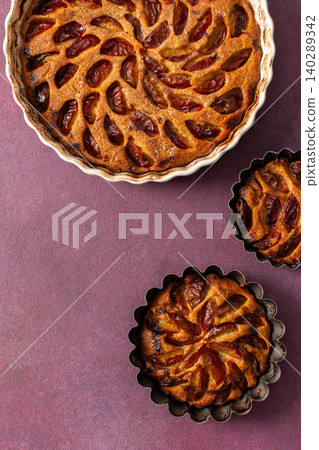 A plum pie is placed on a table with thinly sliced plums visible on top. Bowls of fresh plums are nearby with empty plates waiting for dessert A plum pie is placed on a table with thinly sliced plums visible on top. Bowls of fresh plums are nearby with empty plates waiting for dessert 140289342