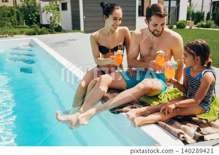 Happy family,rented a house with a swimming pool and enjoy the summer holiday. father mother and daughter sitting at the poolside and drinking orange cocktail Happy family,rented a house with a swimming pool and enjoy the summer holiday. father mother and daughter sitting at the poolside and drinking orange cocktail 140289421