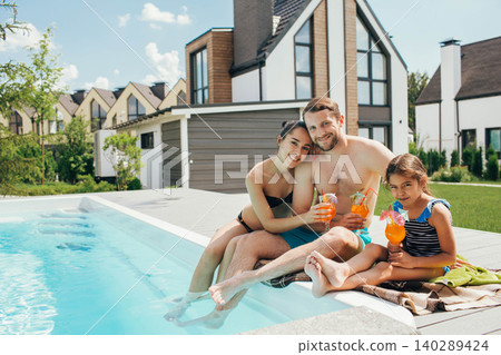 Happy family,rented a house with a swimming pool and enjoy the summer holiday. father mother and daughter sitting at the poolside and drinking orange cocktail Happy family,rented a house with a swimming pool and enjoy the summer holiday. father mother and daughter sitting at the poolside and drinking orange cocktail 140289424