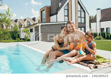 Happy family, summer resting in the pool. father mother and daughter sitting at the poolside and drinking orange cocktail Happy family, summer resting in the pool. father mother and daughter sitting at the poolside and drinking orange cocktail 140289425