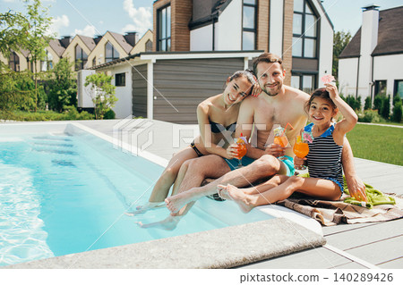 Happy family,rented a house with a swimming pool and enjoy the summer holiday. father mother and daughter sitting at the poolside and drinking orange cocktail Happy family,rented a house with a swimming pool and enjoy the summer holiday. father mother and daughter sitting at the poolside and drinking orange cocktail 140289426