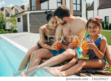 Happy family,rented a house with a swimming pool and enjoy the summer holiday. father mother and daughter sitting at the poolside and drinking cocktail Happy family,rented a house with a swimming pool and enjoy the summer holiday. father mother and daughter sitting at the poolside and drinking cocktail 140289427