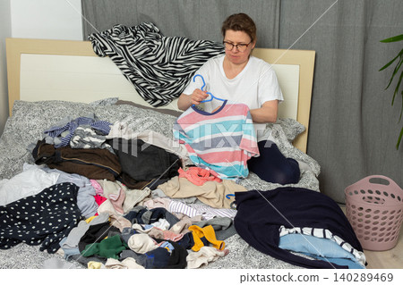 A 52-year-old woman sorts laundry on a bed. She holds a shirt and organizes various garments scattered around. A laundry basket sits nearby as she works on the task at hand A 52-year-old woman sorts laundry on a bed. She holds a shirt and organizes various garments scattered around. A laundry basket sits nearby as she works on the task at hand 140289469