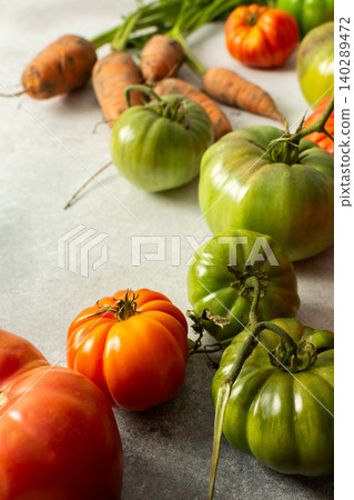 Various types of tomatoes and carrots are placed together on a flat surface. The assortment shows green and ripe tomatoes along with leafy greens and roots Various types of tomatoes and carrots are placed together on a flat surface. The assortment shows green and ripe tomatoes along with leafy greens and roots 140289472