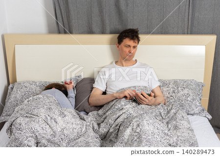 A married wakes up in bed at home. One partner sits up looking at a phone while the other sleeps. They are surrounded by blankets and pillows in a peaceful room A married wakes up in bed at home. One partner sits up looking at a phone while the other sleeps. They are surrounded by blankets and pillows in a peaceful room 140289473