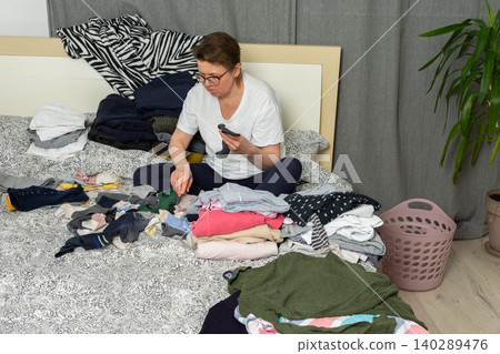 A 52-year-old woman sits on her bed, sorting a pile of laundry. Clothes are spread out around her as she carefully organizes socks and other garments in her bedroom A 52-year-old woman sits on her bed, sorting a pile of laundry. Clothes are spread out around her as she carefully organizes socks and other garments in her bedroom 140289476