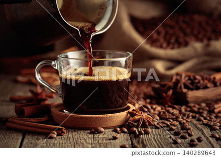 Black coffee is poured from a copper coffee pot into a glass cup. Black coffee is poured from a copper coffee pot into a glass cup. 140289643
