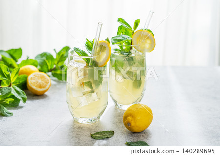 A summer drink with lemon, mint, and ice in a two glasses against a sunny window background. A summer drink with lemon, mint, and ice in a two glasses against a sunny window background. 140289695