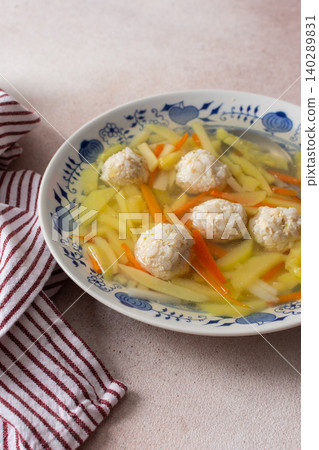 A bowl filled with vegetable soup featuring chicken meatballs sits on a surface with a striped cloth nearby. This dish is ideal for any meal during the day A bowl filled with vegetable soup featuring chicken meatballs sits on a surface with a striped cloth nearby. This dish is ideal for any meal during the day 140289831
