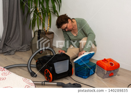 A person is getting ready to vacuum a room. They are attaching a container to the vacuum cleaner while kneeling on the floor. A plant is visible in the background A person is getting ready to vacuum a room. They are attaching a container to the vacuum cleaner while kneeling on the floor. A plant is visible in the background 140289835