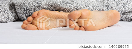 A man enjoys his time relaxing on a bed. His feet can be seen sticking out from under a gray blanket. It looks like a comfortable setting at home or in a hotel, banner 140289955