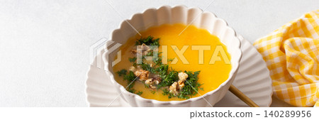 A bowl of pumpkin cream soup sits on a plate. The soup contains nuts and herbs. A yellow checkered napkin lies nearby. This meal can be enjoyed at breakfast or lunch, banner A bowl of pumpkin cream soup sits on a plate. The soup contains nuts and herbs. A yellow checkered napkin lies nearby. This meal can be enjoyed at breakfast or lunch, banner 140289956