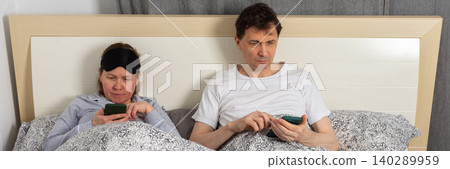 A fifty-year-old wakes up in their bedroom. Both are in bed with blankets. They are looking at their phones in the quiet of the morning, enjoying a relaxed start to the day, banner A fifty-year-old wakes up in their bedroom. Both are in bed with blankets. They are looking at their phones in the quiet of the morning, enjoying a relaxed start to the day, banner 140289959