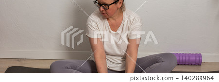A 52-year-old woman exercises at home, sitting on a yoga mat. She is stretching and holding her feet while focusing on her breathing and posture in a simple indoor setting, banner A 52-year-old woman exercises at home, sitting on a yoga mat. She is stretching and holding her feet while focusing on her breathing and posture in a simple indoor setting, banner 140289964