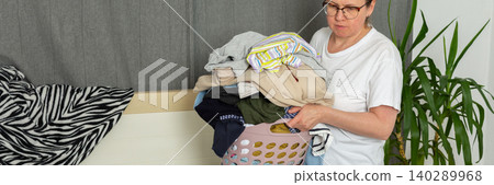 A 52-year-old woman pours laundry out of a basket onto her bed. She sorts through the clothes in her comfortable room as part of her routine tasks, banner A 52-year-old woman pours laundry out of a basket onto her bed. She sorts through the clothes in her comfortable room as part of her routine tasks, banner 140289968