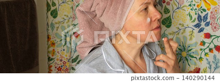 A 50-year-old woman prepares her skin by applying cream in her bathroom. She wears a towel on her head and stands in front of a colorful shower curtain during her morning routine, banner A 50-year-old woman prepares her skin by applying cream in her bathroom. She wears a towel on her head and stands in front of a colorful shower curtain during her morning routine, banner 140290144