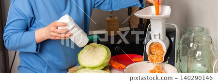 A woman is working in a kitchen to prepare fermented cabbage. She is using a grater and mixing in shredded carrots while measuring salt. The kitchen has different utensils and jars, banner A woman is working in a kitchen to prepare fermented cabbage. She is using a grater and mixing in shredded carrots while measuring salt. The kitchen has different utensils and jars, banner 140290146