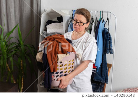 A person stands in a room holding a basket full of dirty clothes. Garments hang in the background. The setting is bright and casual, focusing on the laundry task ahead A person stands in a room holding a basket full of dirty clothes. Garments hang in the background. The setting is bright and casual, focusing on the laundry task ahead 140290148