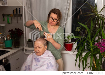 A woman uses a comb to cut her mother hair in the kitchen. They share a special moment together. The room is bright with plants nearby and tools hung on the wall A woman uses a comb to cut her mother hair in the kitchen. They share a special moment together. The room is bright with plants nearby and tools hung on the wall 140290149