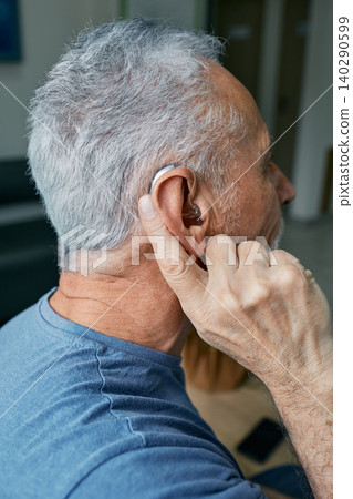Senior man tunes his hearing aid behind the ear by pressing his finger on setting button. Modern hearing aid, hearing solutions Senior man tunes his hearing aid behind the ear by pressing his finger on setting button. Modern hearing aid, hearing solutions 140290599