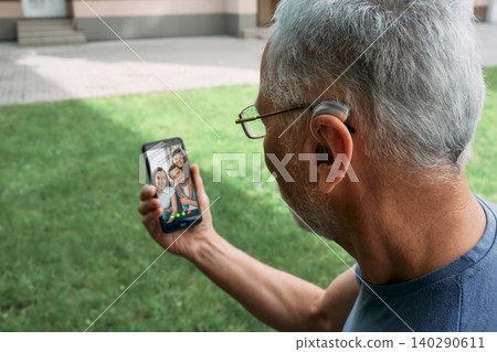 Senior man with a hearing aid behind the ear communicates with his family via video communication via a smartphone. Full human life with hearing aids Senior man with a hearing aid behind the ear communicates with his family via video communication via a smartphone. Full human life with hearing aids 140290611