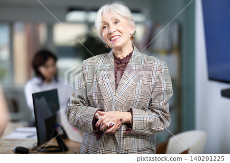 Senior woman employee stands in buzzing office, in middle of working day Senior woman employee stands in buzzing office, in middle of working day 140291225