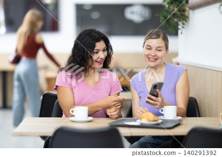 Latin girl is chatting with female friend at table in cafe, showing photos on phone screen 140291392