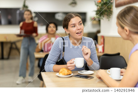 Young girl chatting with friend over coffee and croissant in cafeteria Young girl chatting with friend over coffee and croissant in cafeteria 140291946
