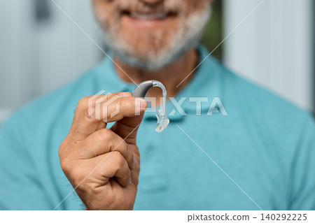 Senior man holding BTE hearing aid in hand on foreground, close-up. Treatment of deafness in elderly people Senior man holding BTE hearing aid in hand on foreground, close-up. Treatment of deafness in elderly people 140292225