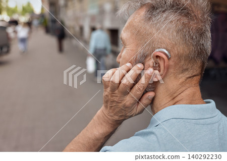 Elderly man with hearing aid behind the ear holds hand near his ear for listening environment urban street, having full life Elderly man with hearing aid behind the ear holds hand near his ear for listening environment urban street, having full life 140292230