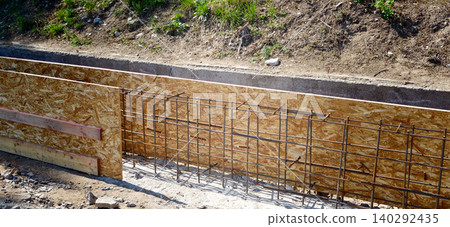 Construction site featuring reinforced concrete fencing with wooden forms and exposed rebar, set against a grassy hillside under clear blue skies Construction site featuring reinforced concrete fencing with wooden forms and exposed rebar, set against a grassy hillside under clear blue skies 140292435