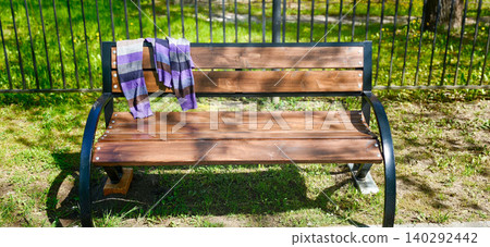 Forgotten sweater draped over a wooden park bench surrounded by green grass and a black fence in a sunny outdoor setting Forgotten sweater draped over a wooden park bench surrounded by green grass and a black fence in a sunny outdoor setting 140292442