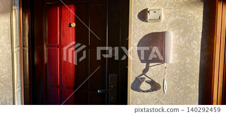 Entrance door with intercom system in an apartment hallway, illuminated by warm sunlight casting shadows on the textured wall Entrance door with intercom system in an apartment hallway, illuminated by warm sunlight casting shadows on the textured wall 140292459