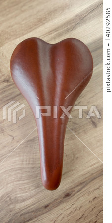 Bicycle seat with a heart-shaped design, crafted from brown leather, placed on a wooden surface showcasing natural grain patterns and textures Bicycle seat with a heart-shaped design, crafted from brown leather, placed on a wooden surface showcasing natural grain patterns and textures 140292585