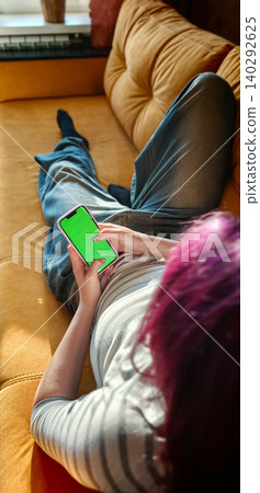 Young woman with purple hair lying on a yellow sofa, holding a smartphone with a green screen, relaxed indoor setting with natural light streaming through a window Young woman with purple hair lying on a yellow sofa, holding a smartphone with a green screen, relaxed indoor setting with natural light streaming through a window 140292625