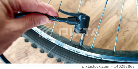 Hand holding bicycle pump nozzle to inflate bicycle tire valve, close-up view of wheel rim and spokes on wooden surface, focus on pump connection Hand holding bicycle pump nozzle to inflate bicycle tire valve, close-up view of wheel rim and spokes on wooden surface, focus on pump connection 140292862
