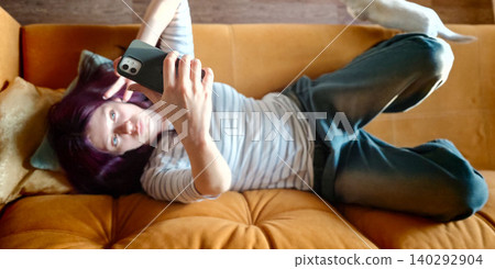 Young woman with purple hair lying on a brown sofa, taking a selfie with a smartphone, casual clothing and decorative pillows visible in a cozy indoor setting Young woman with purple hair lying on a brown sofa, taking a selfie with a smartphone, casual clothing and decorative pillows visible in a cozy indoor setting 140292904