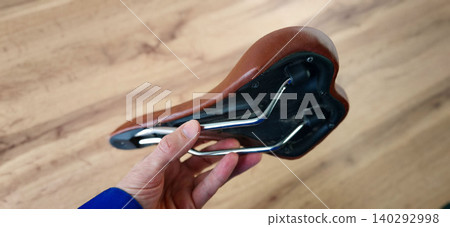 Bicycle seat held in hand, showcasing brown leather material and metal frame, placed on a wooden surface with visible grain patterns and textures Bicycle seat held in hand, showcasing brown leather material and metal frame, placed on a wooden surface with visible grain patterns and textures 140292998