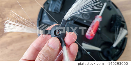 Hand holding a replacement brush for a robot vacuum cleaner, with a screwdriver and vacuum base visible in the background on a wooden surface Hand holding a replacement brush for a robot vacuum cleaner, with a screwdriver and vacuum base visible in the background on a wooden surface 140293000