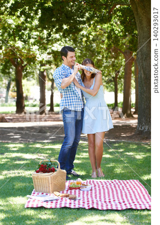 Love, picnic and surprise with couple in park for romance, bonding and summer vacation. Happiness, commitment and relax with man and blindfold woman in nature for date, support and relationship 140293017