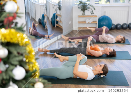 Women practice yoga during the New Year celebration in the studio, lie on the mat, perform stretching in Ardha Pavanmuktasana pose Women practice yoga during the New Year celebration in the studio, lie on the mat, perform stretching in Ardha Pavanmuktasana pose 140293264