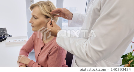 Audiologist putting hearing aid on mature woman's ear at audiology center, close-up, side view. Deafness treatment, hearing solutions Audiologist putting hearing aid on mature woman's ear at audiology center, close-up, side view. Deafness treatment, hearing solutions 140293424
