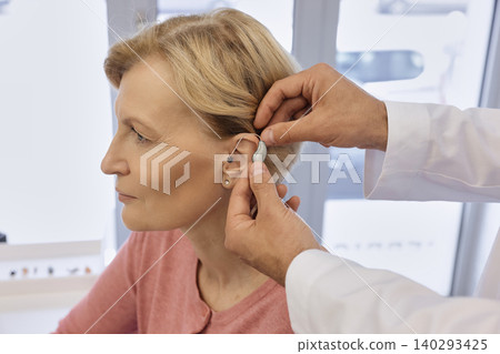 Audiologist installing hearing aid on ear of mature woman in hearing clinic. Deafness Treatment, hearing solutions Audiologist installing hearing aid on ear of mature woman in hearing clinic. Deafness Treatment, hearing solutions 140293425